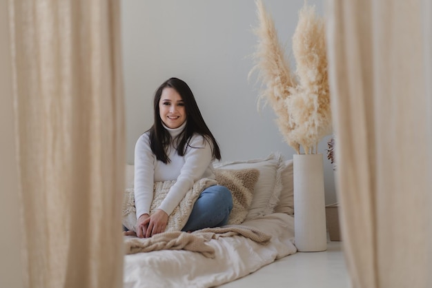 Bella ragazza felice con i capelli lunghi si siede a letto abbracciando un cuscino in interni colori pastello