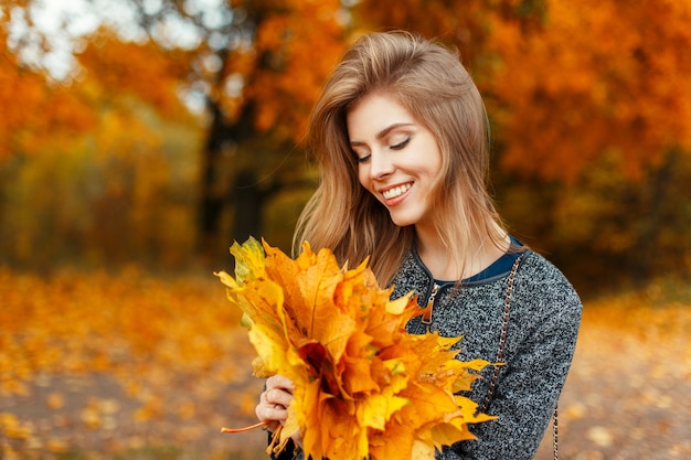 Bella ragazza felice con un sorriso che tiene un mazzo di foglie gialle in un parco in autunno