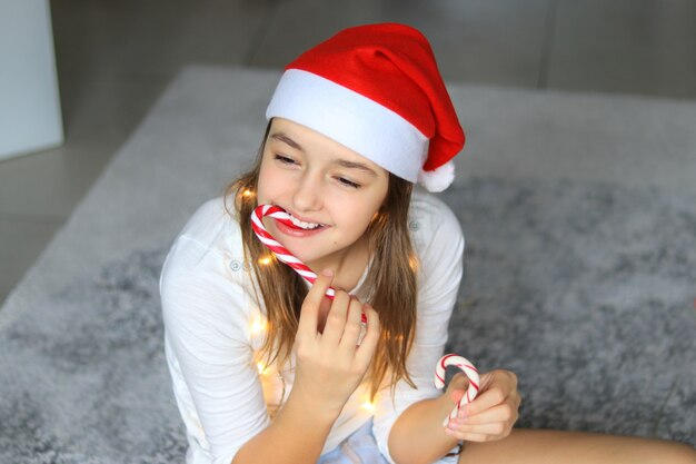 Bella ragazza felice del preteen in cappello rosso del Babbo Natale che mangia i bastoncini di zucchero a strisce di Natale