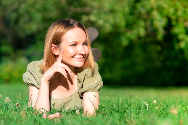 Bella ragazza felice e positiva, giovane donna allegra è sdraiata sul petto sull'erba verde nel parco o nella foresta e sorride, si diverte a camminare, bel tempo caldo e soleggiato, distogliendo lo sguardo, copia spazio