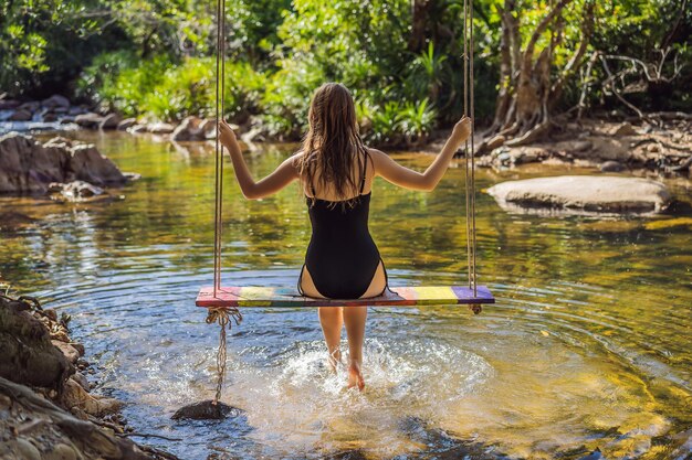 Bella ragazza felice giovane donna abbastanza allegra viaggiatore seduto oscillante su un'altalena legata a un albero sopra l'acqua sorridente in una giornata di sole Vacanze estive concetto di viaggio