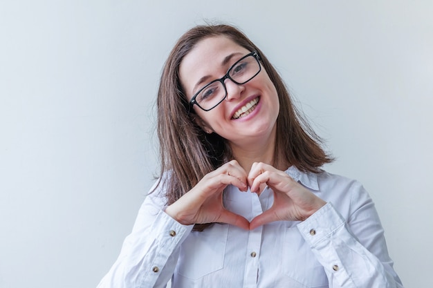 Bella ragazza felice sorridente. Giovane donna semplice del ritratto di bellezza in occhiali che mostrano il segno del cuore con le mani isolate su bianco. Emozioni umane positive espressione facciale linguaggio del corpo.