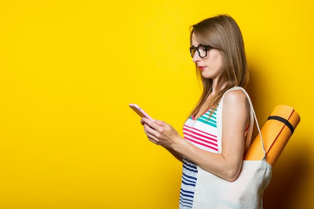 Bella ragazza giovane con karimat in una borsa guarda il telefono su uno sfondo giallo.