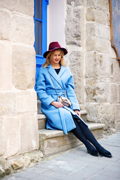 Bella ragazza giovane turista in un cappotto blu e cappello bordeaux seduto sulle scale del bar accogliente con porte blu