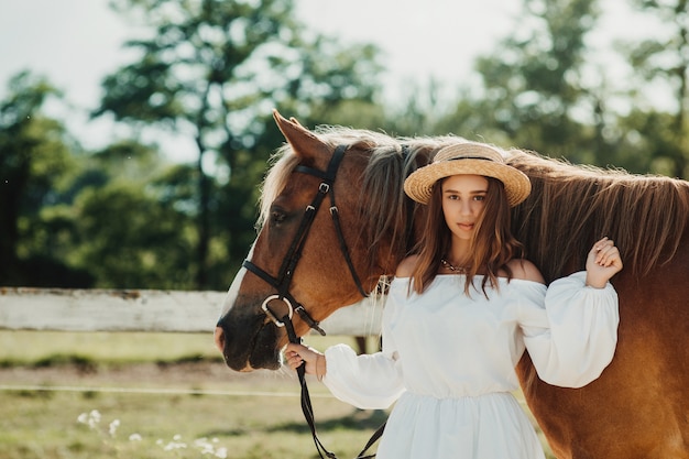 Bella ragazza hippie con cavallo su uno sfondo di ranch, vista frontale. La ragazza con il cappello.