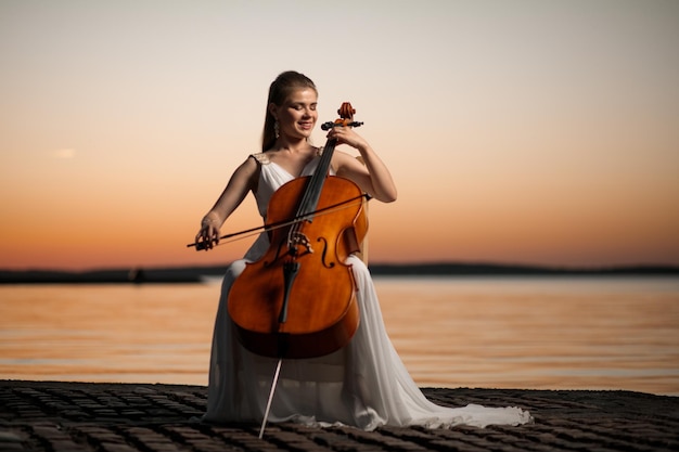 Bella ragazza in abito bianco che suona il violoncello sul lago dopo il tramonto