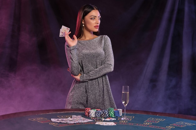 Bella ragazza in abito grigio mostra due carte mentre posa al tavolo da gioco nel casinò Sfondo fumo nero con retroilluminazione colorata Intrattenimento gioco d'azzardo poker champagne Primo piano