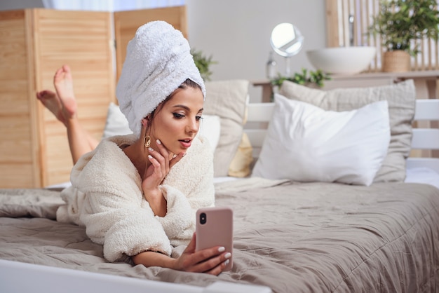 Bella ragazza in accappatoio e con l'asciugamano sulla sua testa che fa selfie facendo uso dello smartphone sul letto