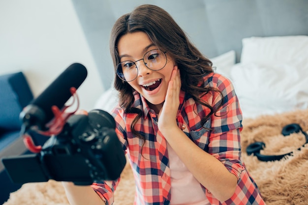 Bella ragazza in camicia a scacchi che indossa occhiali e si tocca la guancia mentre si registra su una telecamera