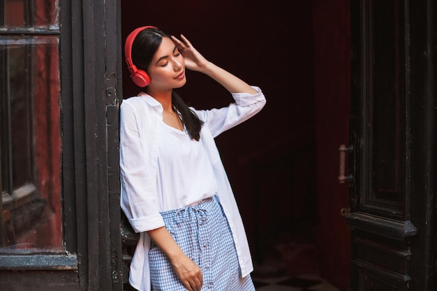 Bella ragazza in camicia bianca che chiude sognante gli occhi appoggiandosi alla vecchia porta mentre ascolta musica in cuffie rosse