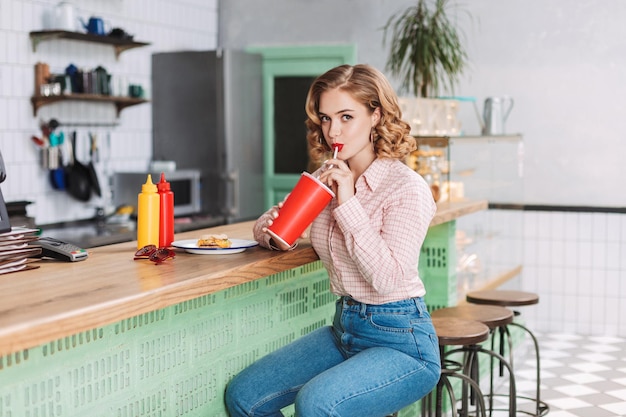 Bella ragazza in camicia e jeans seduta al bancone del bar nella caffetteria e bere acqua gassata mentre guarda tristemente a porte chiuse
