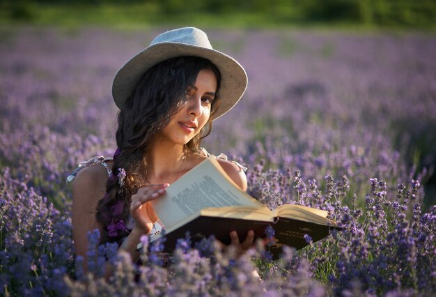 Bella ragazza in cappello che si siede nel campo di lavanda viola e leggendo un libro.