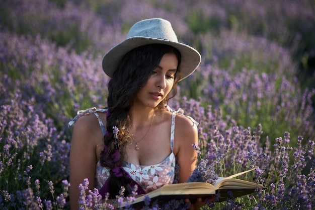 Bella ragazza in cappello che si siede nel campo di lavanda viola e leggere il libro.