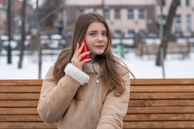 Bella ragazza in cappotto di pelle di pecora si siede su una panchina nel parco invernale e parla sullo smartphone Giovane donna seria con gadget