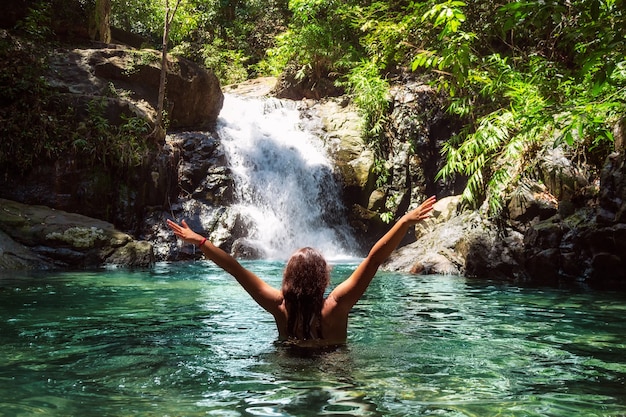 Bella ragazza in costume da bagno con le braccia aperte vicino a una cascata in una foresta tropicale.