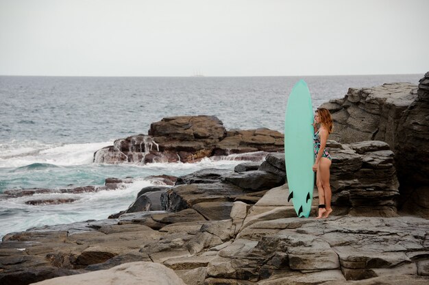 Bella ragazza in costume da bagno multicolore in piedi con la tavola da surf sulla spiaggia rocciosa dell'Oceano Atlantico