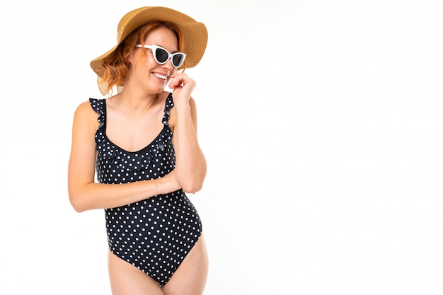 Bella ragazza in costume da bagno retrò e un cappello estivo di paglia e occhiali da sole su un muro bianco con spazio di copia