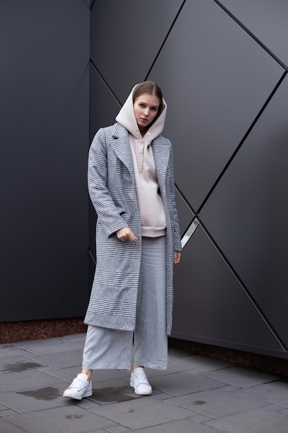 Bella ragazza in elegante cappotto grigio con cappuccio biege alla moda e pantaloni su sfondo argento scuro Camminando per strada