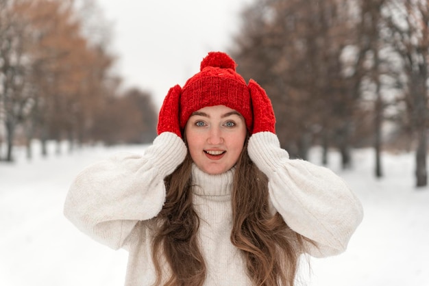 Bella ragazza in lana cappello rosso e guanti sorride direttamente nella fotocamera Donna in abiti a maglia nella foresta invernale