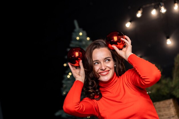 Bella ragazza in maglione rosso con due palline di decorazione vicino alla testa Donna felice davanti all'albero di Natale Bella signora con le palle di Natale sullo sfondo scuro