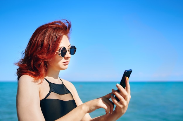 Bella ragazza in occhiali da sole scuri e costume da bagno nero con lo smartphone in mano soleggiata giornata estiva redha...