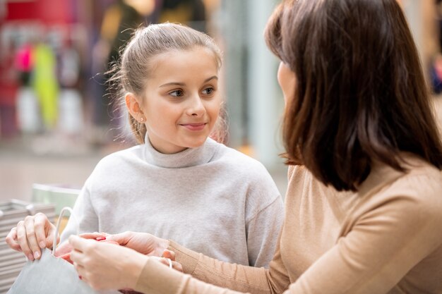 Bella ragazza in pullover bianco che guarda la mamma mentre discute di cosa hanno comprato in vendita venerdì nero nel centro commerciale