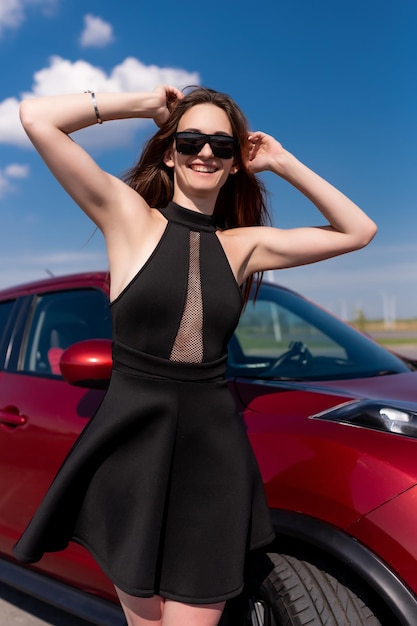 Bella ragazza in un abito corto nero e occhiali da sole si trova vicino a un'auto rossa che tiene i capelli con le mani e sorride