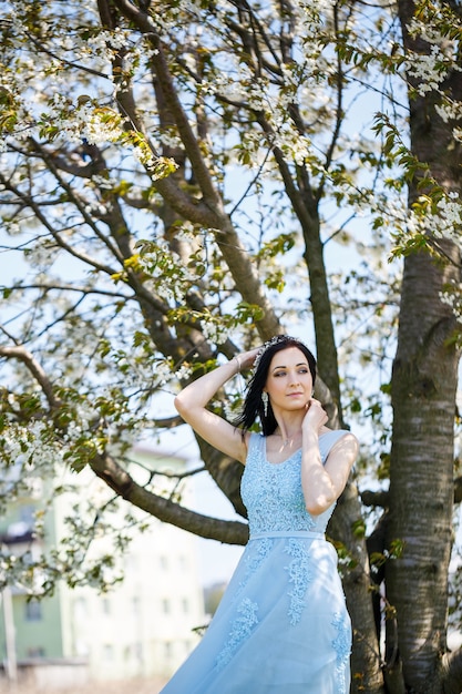 Bella ragazza in un abito lungo blu estivo leggero adornato tra i capelli contro un albero in fiore. Tenero ritratto di una giovane donna in fiore bianco