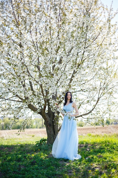 Bella ragazza in un abito lungo blu estivo leggero adornato tra i capelli contro un albero in fiore. Tenero ritratto di una giovane donna in fiore bianco