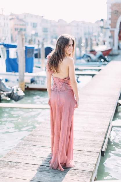 Bella ragazza in un abito rosa in piedi su un ponte di legno tra il canale veneziano e godersi la città.