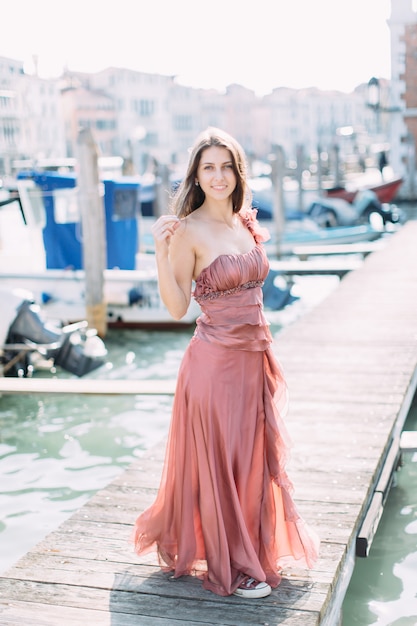 Bella ragazza in un abito rosa in piedi su un ponte di legno tra il canale veneziano e godersi la città.