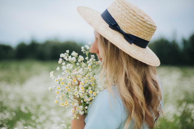 bella ragazza in un campo di margherite. ragazza con un cappello di paglia e un vestito blu. campo di camomilla in estate