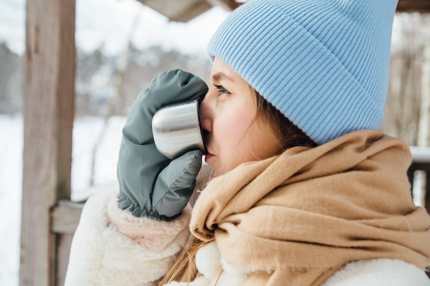 Bella ragazza in un cappotto di pelliccia bianca beve tè caldo in inverno in un bosco innevato