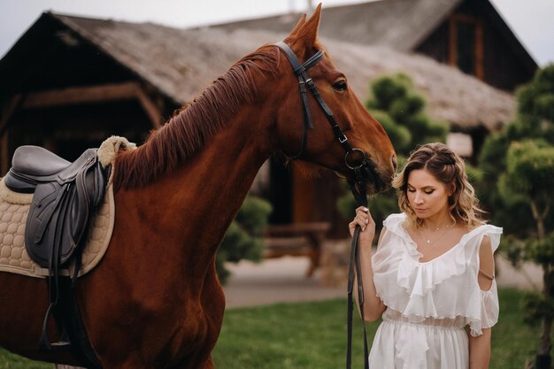 Bella ragazza in un prendisole bianco accanto a un cavallo in un vecchio ranch