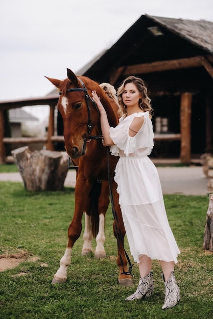 Bella ragazza in un prendisole bianco accanto a un cavallo in un vecchio ranch