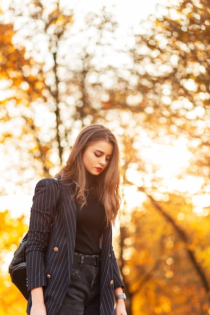 Bella ragazza in un tailleur alla moda con un blazer elegante e un maglione con uno zaino alla moda passeggiate nel parco con fogliame autunnale dorato al tramonto