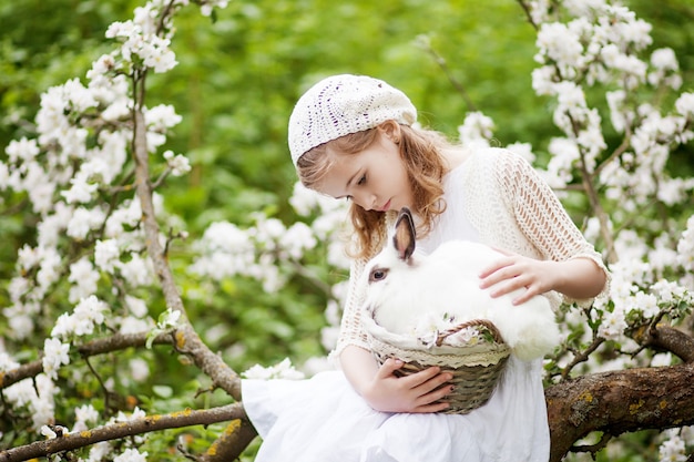 Bella ragazza in un vestito bianco che gioca con il coniglio bianco nel giardino del fiore di primavera. Attività divertente primaverile per bambini. Tempo di Pasqua