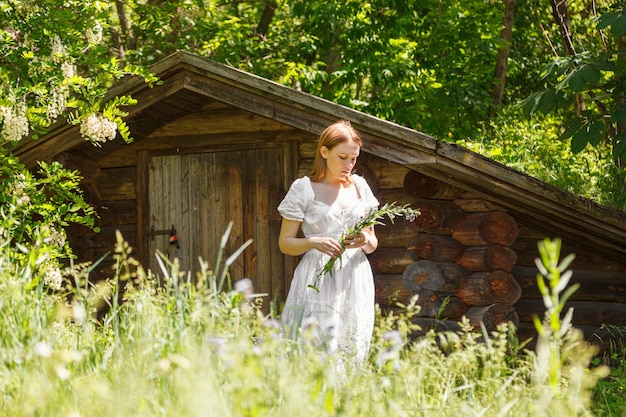 Bella ragazza in un vestito bianco con fiori vicino a una casa di legno