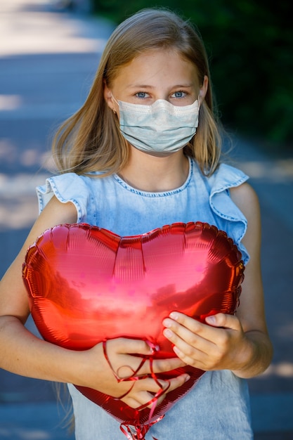 Bella ragazza in un vestito blu con un palloncino a forma di cuore in una maschera medica sul viso