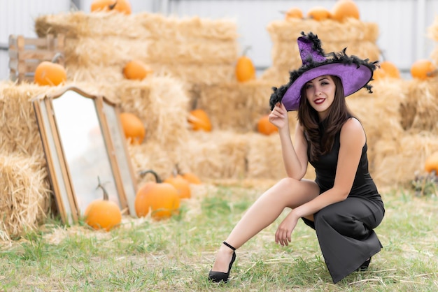 Bella ragazza in un vestito nero e un cappello da strega posa vicino a un vecchio specchio sullo sfondo del fieno decorazioni di zucca di Halloween