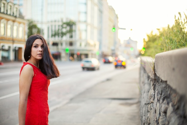 Bella ragazza in un vestito rosso è in piedi vicino alla carreggiata della città, guardando indietro.