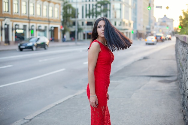 Bella ragazza in un vestito rosso è in piedi vicino alla carreggiata della città, guardando indietro.