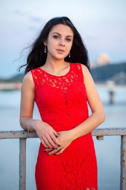 Bella ragazza in un vestito rosso in posa su un ponte vicino a un vecchio recinto. Sullo sfondo c'è un fiume, ponti e frammenti di edifici della città nella sfocatura.