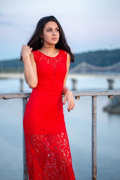 Bella ragazza in un vestito rosso in posa su un ponte vicino a un vecchio recinto. Sullo sfondo c'è un fiume, ponti e frammenti di edifici della città nella sfocatura.