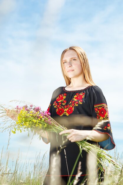 Bella ragazza in un vyshyvanka nero con fiori in un campo che domina il cielo