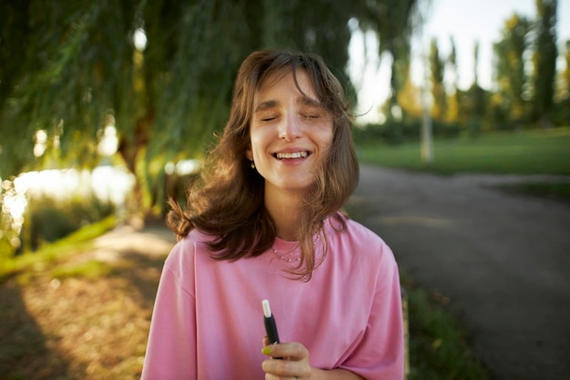 Bella ragazza in una maglietta rosa, sorriso raggiante, con in mano un dispositivo elettronico. Ragazza che usa una sigaretta elettronica.