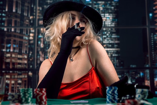 Bella ragazza in vestito lucido rosso e cappello che gioca a poker al casinò