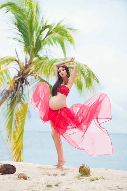 Bella ragazza incinta in vestito rosso sulla spiaggia sabbiosa con le palme