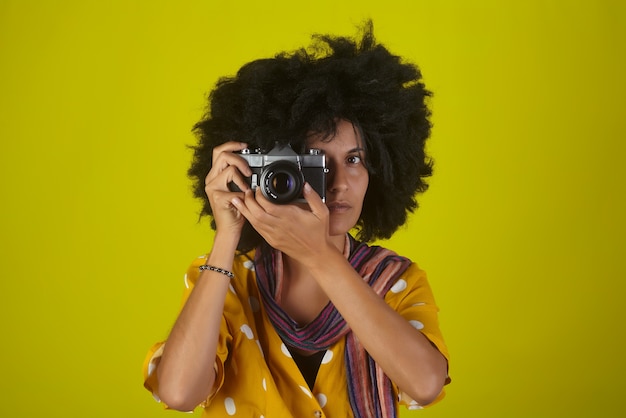 Bella ragazza indiana con acconciatura riccia afro parlando immagini con fotocamera retrò sulla parete gialla