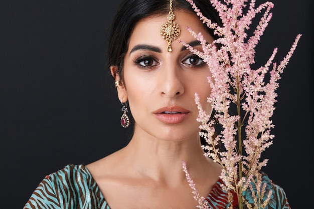 bella ragazza indù con il trucco che indossa il tradizionale abito sari indiano e gioielli etnici che tengono un ramo di fiori isolato su un muro nero